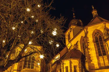Eine historische Kirche bei nächtlicher Beleuchtung. Ein Baum mit leuchtenden Sternen verziert die Szene.