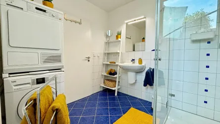 Ein modernes Badezimmer mit blauen Fliesen und einem Duschbereich. Es gibt eine Waschmaschine, einen Handtuchhalter und Pflanzen auf einem Regal.