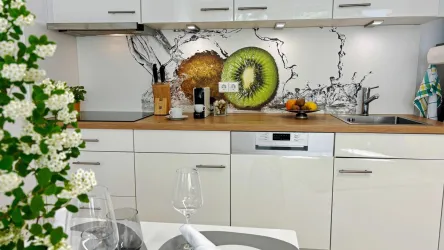Eine moderne Küche mit einem Holzarbeitsplatz und hellen Schränken. Im Hintergrund sind große Fruchtmotive sowie frisches Obst auf dem Tisch sichtbar.