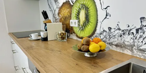 Eine moderne Küche mit Holzarbeitsplatte. Auf dem Tisch steht eine Obstschale mit verschiedenen Früchten und im Hintergrund ein fruchtiges Wanddesign.