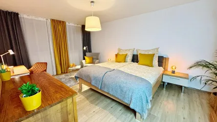 Ein modernes Schlafzimmer mit einem Doppelbett und bunten Kissen. Der Raum ist hell und gemütlich, mit einem Schreibtisch und Pflanzen.