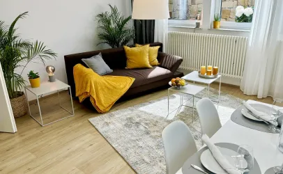 Ein stilvolles Wohnzimmer mit einem braunen Sofa, gelben Kissen und einer Decke. Der Raum hat große Fenster, einen Teppich und eine Esstischgruppe.