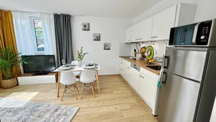 Eine moderne Küche mit einem Essbereich und einem großen Fenster. Der Raum ist hell und freundlich, mit Pflanzen und stilvollen Möbeln.