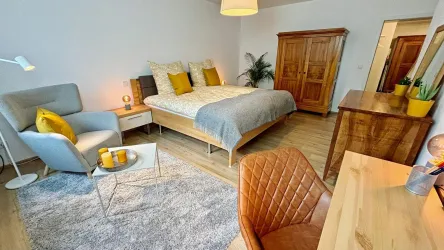 Ein helles Schlafzimmer mit einem großen Bett und gelben Kissen. Es gibt einen gemütlichen Sessel, einen Tisch und eine Holzkommode im Raum.