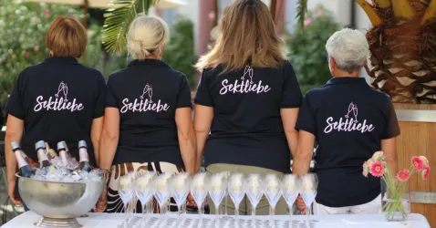 Vier Frauen stehen Rücken an Rücken, tragen schwarze T-Shirts mit der Aufschrift „Sektliebe“. Vor ihnen sind eine Sektschale und viele Gläser auf einem Tisch zu sehen.