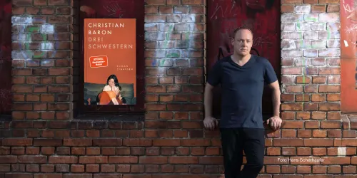 Ein Mann steht an einer Backsteinwand neben einem Plakat mit dem Titel "Drei Schwestern" von Christian Baron. Die Umgebung wirkt urban und künstlerisch.