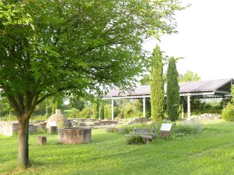 Ein ruhiger Park mit einer großen grünen Wiese und mehreren Bäumen. Im Hintergrund sind Überreste von Mauern und eine überdachte Fläche zu sehen.
