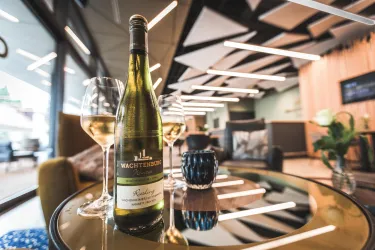 Ein Glas Weißwein steht neben einer Flasche Wachtelburg Riesling auf einem Tisch. Im Hintergrund sind elegante Möbel und eine moderne Einrichtung zu sehen.