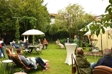 Eine Gartenveranstaltung mit mehreren Menschen, die in Stühlen sitzen und zuhören. Im Hintergrund sind Sonnenschirme und Bäume zu sehen.