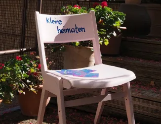 Ein weißer Stuhl mit der Aufschrift „Kleine heimat“ steht vor blühenden Pflanzen. Auf dem Stuhl liegen einige bunte Flyer.