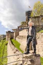Eine Person steht auf einer alten Burgmauer und zeigt mit dem Finger auf etwas im Hintergrund. Die Burg ist von einer grünen Wiese umgeben und hat eine beeindruckende Architektur.