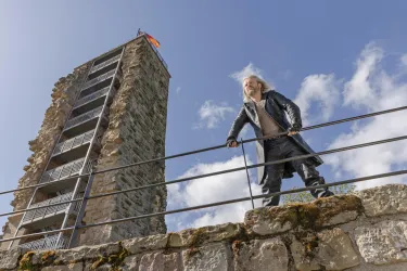 Ein Mann steht auf einer Mauer und blickt auf einen Steinturm. Im Hintergrund sind blaue Himmel und einige Wolken zu sehen.