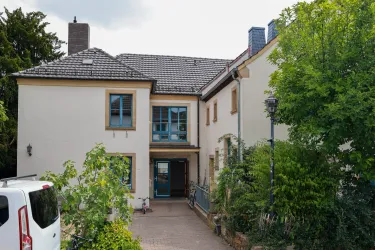 Ein modernes Haus mit einer hellen Fassade und blauen Fenstern. Der Eingangsbereich ist von Pflanzen und Bäumen umgeben.