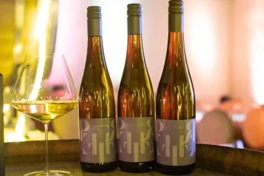 Drei Flaschen Wein stehen auf einem Holzfass, daneben ein Glas mit Weißwein. Die Umgebung ist warm beleuchtet und schafft eine einladende Atmosphäre.