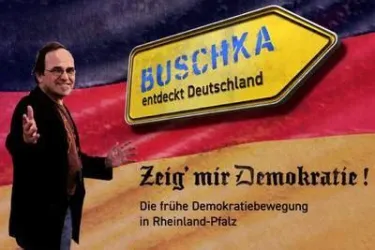 Ein Mann steht vor einem großen Schild mit der Aufschrift "BUSCHKA entdeckt Deutschland". Im Hintergrund ist die deutsche Flagge zu sehen, und der Text fordert auf: "Zeig' mir Demokratie!"