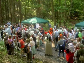 Eine Gruppe von Menschen versammelt sich in einem Wald, umgeben von Bäumen. Es gibt zwei grüne Zelte, und die Atmosphäre wirkt gesellig und einladend.
