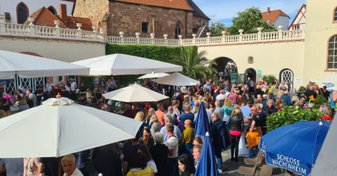 Eine große Menschenmenge versammelt sich in einem Innenhof mit Sonnenschirmen. Im Hintergrund sind historische Gebäude und grüne Pflanzen zu sehen.