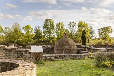 Eine historische Ruine mit Steinstrukturen und einer Kuppel. Umgeben von Bäumen und einem grünen Rasen unter einem hellblauen Himmel.