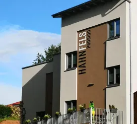 Ein modernes Gebäude mit der Aufschrift "HAHN FELS Pension". Die Fassade ist in hellen Farben gehalten und hat große Fenster.