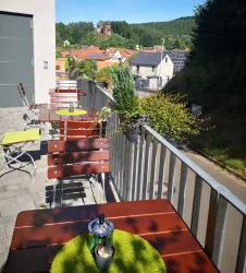 Ein gemütlicher Außenbereich mit Holzttischen und grünen Platzdeckchen. Im Hintergrund ist eine malerische Landschaft mit Hügeln und Häusern zu sehen.