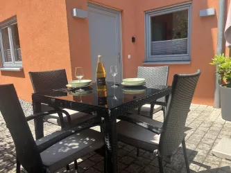 Ein eleganter Tisch mit sechs Stühlen steht auf einem gepflasterten Boden. Auf dem Tisch befinden sich Gläser, Teller und eine Flasche Sekt.