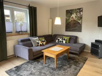 Ein modernes Wohnzimmer mit einem grauen Sofa und bunten Kissen. Ein Holztisch und ein großer Teppich ergänzen die gemütliche Einrichtung.