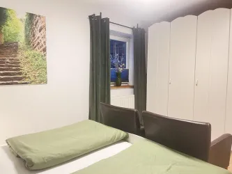 Ein einfaches Schlafzimmer mit zwei Betten und grünen Bettlaken. Im Hintergrund sind ein Kleiderschrank und ein Fenster mit Vorhängen zu sehen.