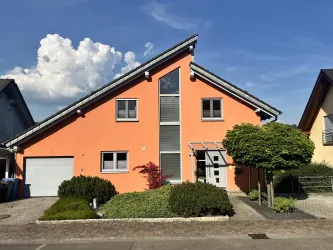 Ein modernes, orangefarbenes Haus mit einem schrägen Dach. Der Vorgarten ist gepflegt und es gibt einige Bäume und Sträucher.