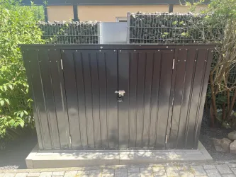Ein schlossgesicherter Geräteschuppen aus Metall steht im Garten. Umgeben von Pflanzen und einer hohen Hecke bietet er Stauraum für Werkzeuge.