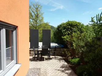 Ein gemütlicher Außenbereich mit einem Sitzplatz und einem Tisch. Umgeben von grünen Pflanzen und einem klaren blauen Himmel.