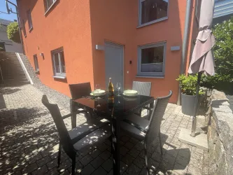 Ein gemütlicher Außenbereich mit einem Tisch und Stühlen. Die orangefarbenen Wände des Hauses und die grünen Pflanzen schaffen eine einladende Atmosphäre.