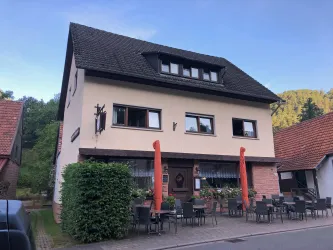 Ein gemütliches Haus mit einer hellen Fassade und einem Garten. Vor dem Gebäude stehen Stühle und Tische zur Draußensitzgelegenheit.