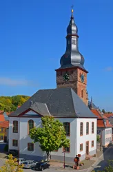 Ein historisches Gebäude mit einem markanten Turm und einer großen Uhr. Der Himmel ist klar und blau, umgeben von schönen Bäumen.