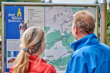 Zwei Personen betrachten eine Wandkarte des Adolf-Kassau-Bänderwegs. Die Karte zeigt Wanderwege und umliegende Gebiete.