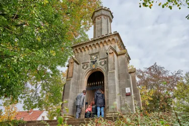 Ein historisches Gebäude umgeben von Bäumen und buntem Herbstlaub. Menschen stehen vor dem Eingang und erkunden die Architektur.