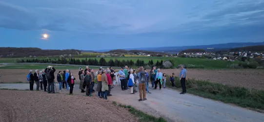 Eine Gruppe von Menschen steht auf einem Feldweg bei Dämmerung. Im Hintergrund sind grüne Felder und ein größeres Dorf sichtbar.