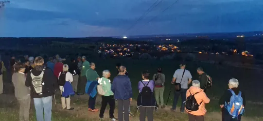 Eine Gruppe von Menschen steht auf einem Hügel und schaut auf ein beleuchtetes Dorf im Abendlicht. Der Himmel ist leicht bewölkt und es ist spät am Tag.