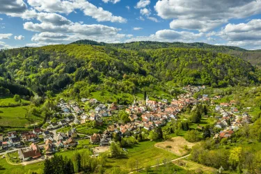 Eine malerische Aussicht auf ein kleines Dorf umgeben von grünen Hügeln und Wäldern. Der Himmel ist blau mit einigen weißen Wolken.