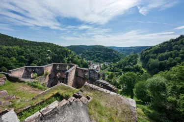Eine alte Burgruine mit Blick auf eine grüne Landschaft und bewaldete Hügel. Der Himmel ist klar und blau mit einigen Wolken.