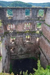 Eine alte Ruine mit offenen Fenstern und zerfallenen Wänden. Umgeben von grünen Hügeln und einem klaren Himmel.