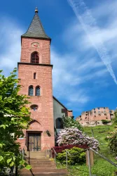 Eine alte Kirche aus roten Steinen mit einem hohen Turm. Im Vordergrund sind Blumen und Bäume zu sehen, bei klarem blauen Himmel.