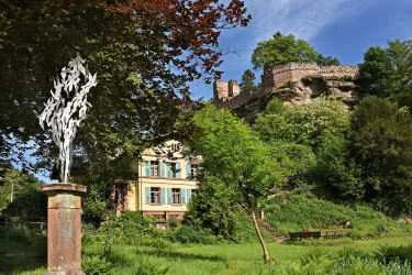 Eine schöne Villa mit einem Kunstwerk im Vordergrund, umgeben von grüner Natur. Im Hintergrund sieht man eine Burgruine auf einem Hügel.