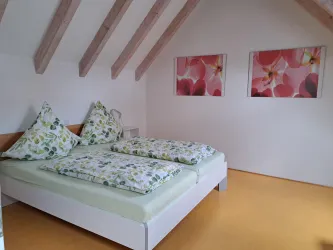 Ein gemütliches Schlafzimmer mit einem Doppelbett und bunten Kissen. An den Wänden hängen Bilder von Blumen.
