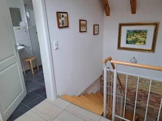 Ein heller Flur mit einer Holztreppe und Bildern an der Wand. Im Hintergrund ist ein Badezimmer sichtbar, das modern gestaltet ist.