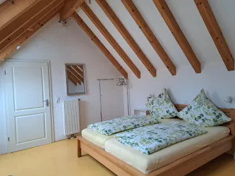 Ein gemütliches Schlafzimmer mit einem Doppelbett und hohen Holzdecken. Durch große Fenster strömt das Licht in den Raum.