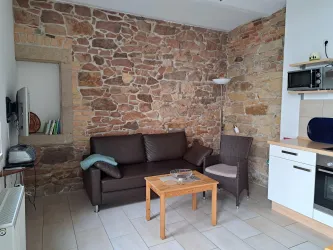 Ein gemütliches Wohnzimmer mit Natursteinwand und einem braunen Sofa. Auf einem Holztisch stehen mehrere Gegenstände und es gibt eine moderne Küchenzeile.