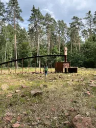 Eine Person geht durch eine lichtdurchflutete Waldlandschaft mit grünen Bäumen. Im Hintergrund steht eine moderne Skulptur aus Metall und Holz.