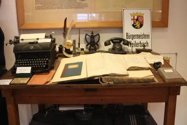 Ein Holzschreibtisch mit einer alten Schreibmaschine, einem Telefon und verschiedenen Dokumenten. Auf dem Tisch liegen auch Bücher und ein Schild mit der Aufschrift "Bürgermeisterei Heilbronn".