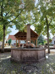 Ein schöner Brunnen steht im Mittelpunkt eines Platzes, umgeben von Bäumen. Im Hintergrund sieht man ein schattiges Pavillon und einige Häuser.