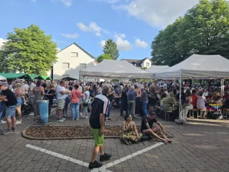 Eine Menschenmenge versammelt sich auf einem Fest mit mehreren weißen Zelten. Im Hintergrund sind grüne Bäume und ein blauer Himmel zu sehen.